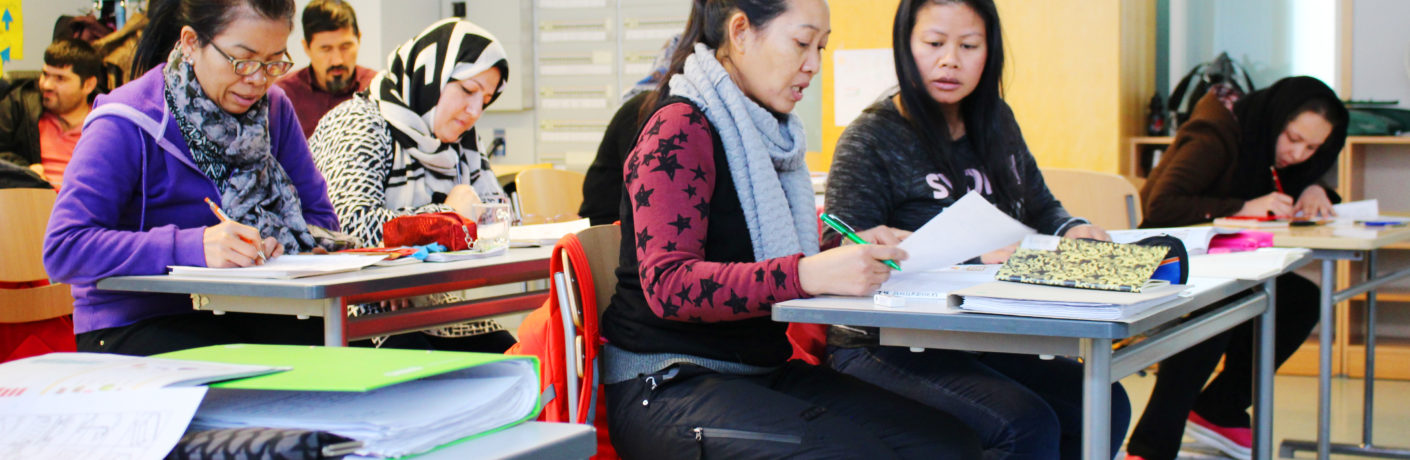 eri maista tulleita naisia ja miehiä istumassa koululuokassa pulpettien äärellä opiskelemassa