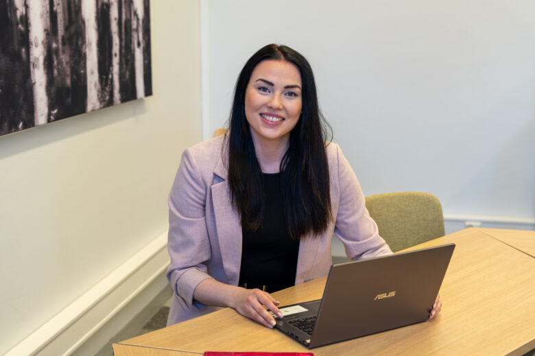 Tummahiuksinen henkilö istuu vihreällä tuolilla kokoushuoneen pöydän äärellä kannettavan tietokoneen äärellä katsoen kameraan.