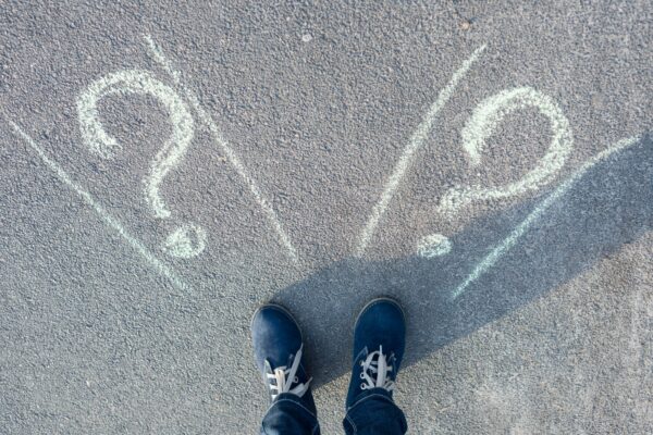 Kuvassa näkyy henkilön jalat, joka seisoo kadulla, johon piirretty kaksi kysymysmerkkiä kuin kaksi eri suuntaa, johon lähteä.