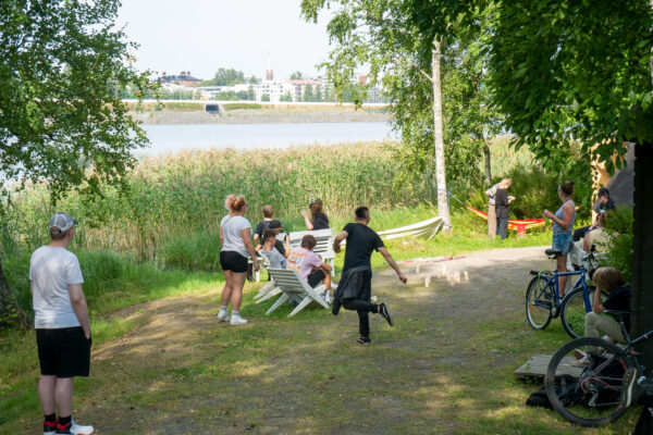 Joukko opiskelijoita pelaamassa aurinkoisessa kesäsäässä mölkkyä järven rannalla,