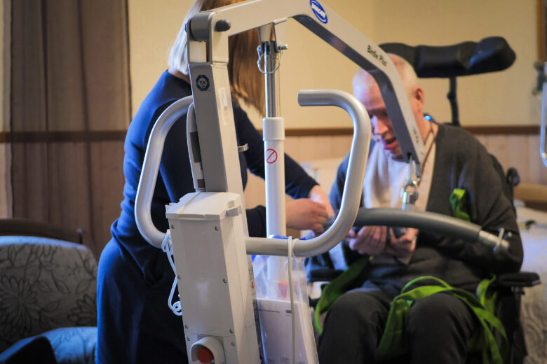 Hoiva-avustaja auttaa henkilöä nostolaitteella pyörätuoliin