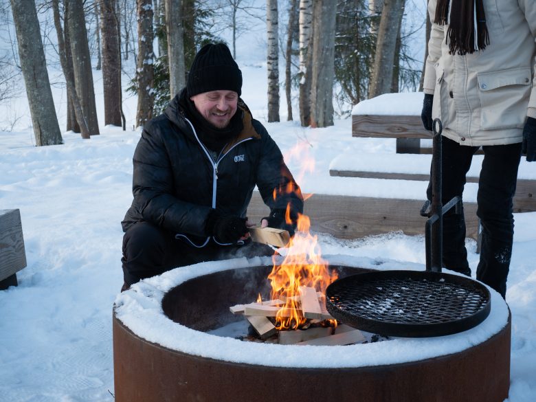 Mies istuu nuotiopaikalla ja lisää puita nuotioon