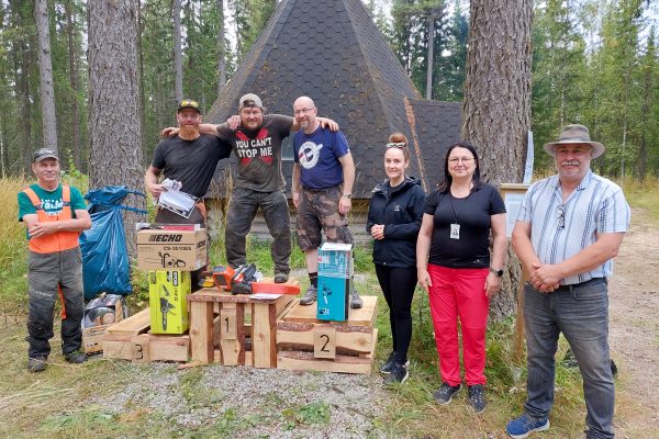 Moottorisahaveistokilpailun voittajat lavoista tehdyillä korokkeilla. Vieressä seisoo kilpailun tuomaristoa ulkoiluvaatteissa. Kuva on otettu metsämaisemassa laavun edustalla.