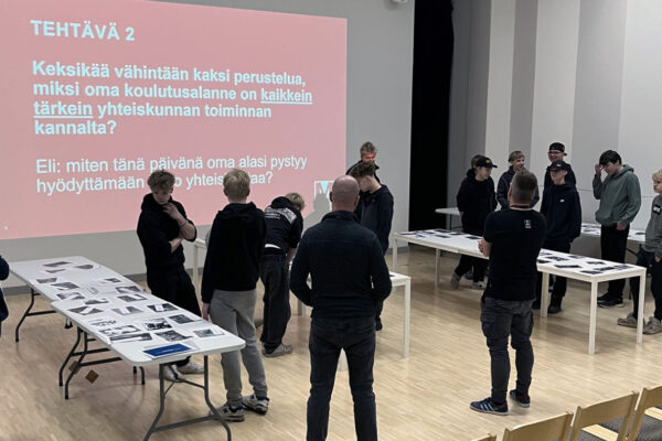 Ihmiset seisovat kahden eri pöydän ympärillä. Taustalla isolla valkokankaalla lukee kysymys: Keksikää vähintään kaksi perustelua miksi oma koulutusalanne on kaikkein tärkein yhteiskunnan toiminnan kannalta