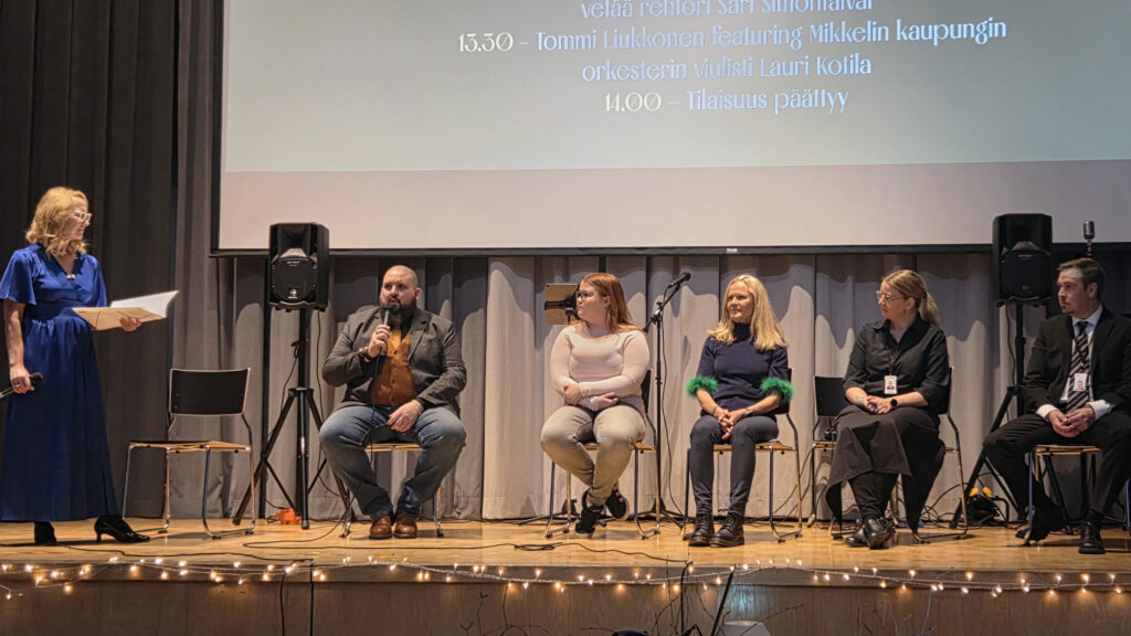 Viisi panelistia istuu tuolissa, paneelikeskustelun juontaja seisoo kuvan vasemmassa reunassa