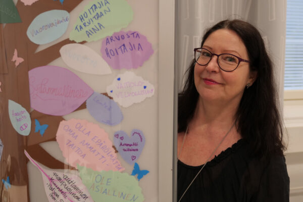 Nainen kurkistaa sermin takaa ja katsoo kameraan hymyillen