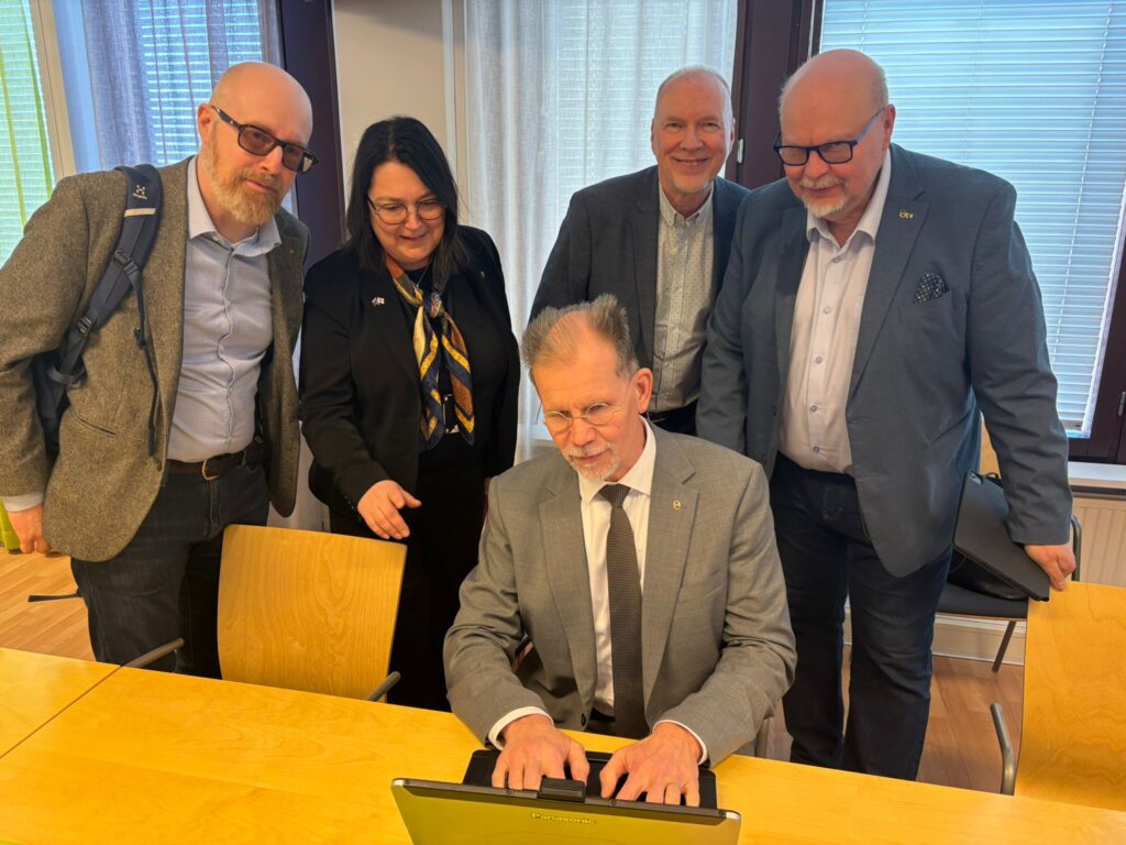 Neljä henkilöä seisoo pöydän ympärillä. Pöydän ääressä istuu yksi henkilö koneen ääressä.