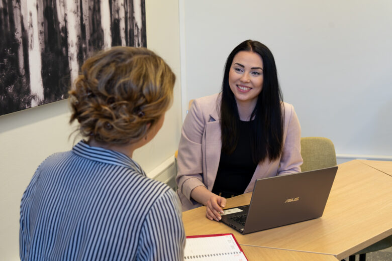 kaksi henkilöä keskustelevat neuvottelutilassa pöydän äärellä kannettavantietokeen ja muistion kanssa. Toinen henkiöistä on raidallinen paita päällä selkä kameraan ja toinen henkilö on kameraanpäin yllään vaalea jakku.