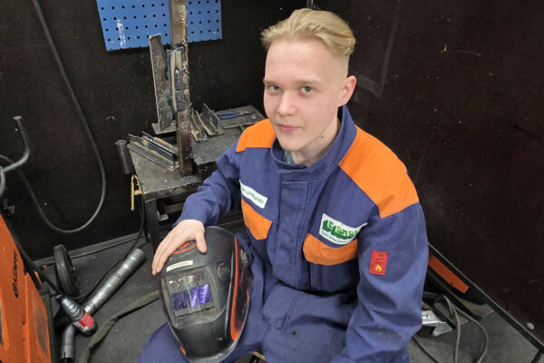 Mies katsoo kameraan hymyillen, pitää sylissä hitsausmaskia.