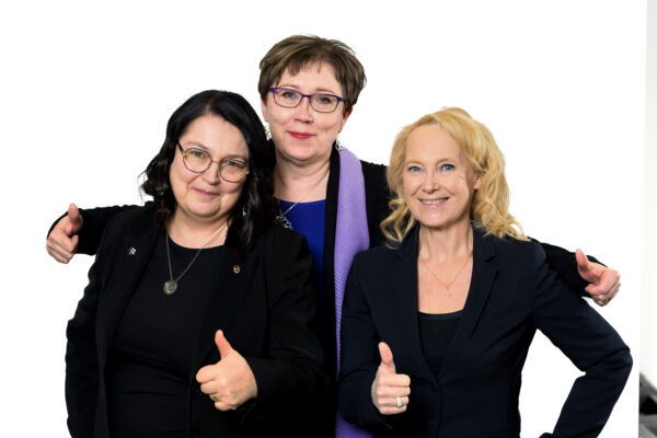 Kolme naista katsoo hymyillen kameraan ja näyttävät peukkua