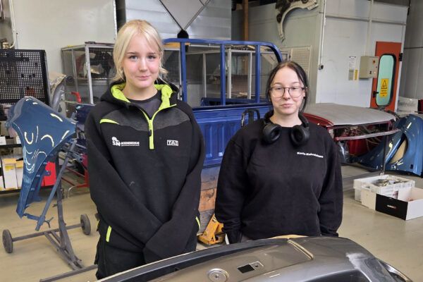Kaksi naista seisoo autokorjaamohallissa katsoen kameraan.