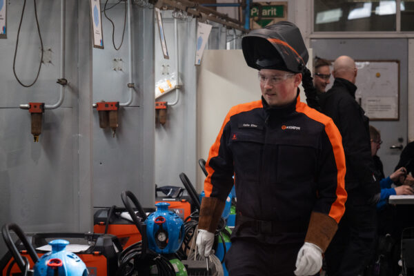 Mies seisoo hitsauskoneiden vieressä työvaatteissa. Taustalla kaksi henkilöhahmoa.