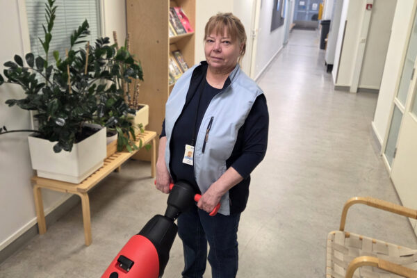 Nainen katsoo kameraan. Seisoo käytävällä, käsissään lattianpesukone